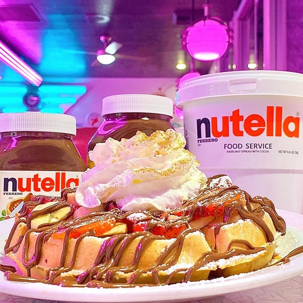 A waffle topped with strawberries, bananas, whipped cream, and Nutella, with Nutella jars in the background.