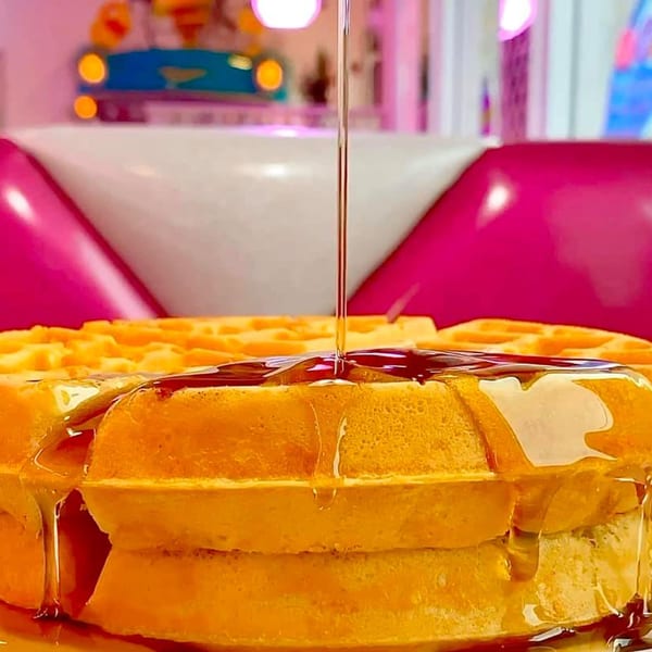 Syrup pouring over a stack of golden waffles at Beeps Diner, with pink and white booths in the background.