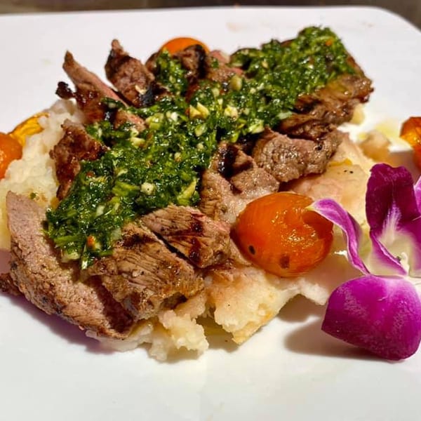 Grilled steak with green chimichurri sauce, roasted tomatoes, and mashed potatoes, garnished with an orchid.