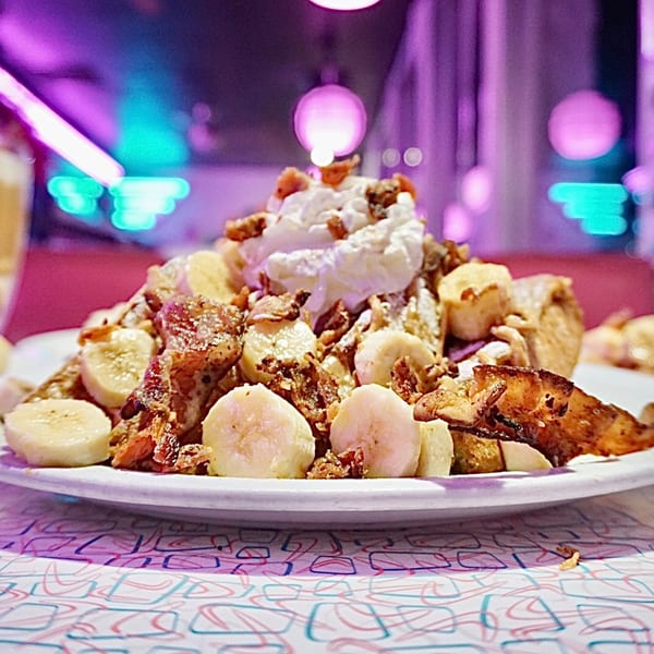 Beeps Diner's French toast with bananas, bacon, whipped cream, and nuts under neon lights.