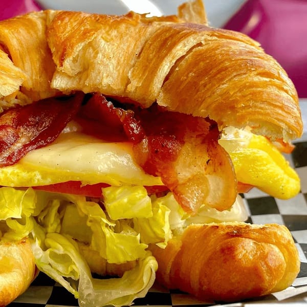 Close-up of a croissant breakfast sandwich with bacon, egg, cheese, tomato, and lettuce.