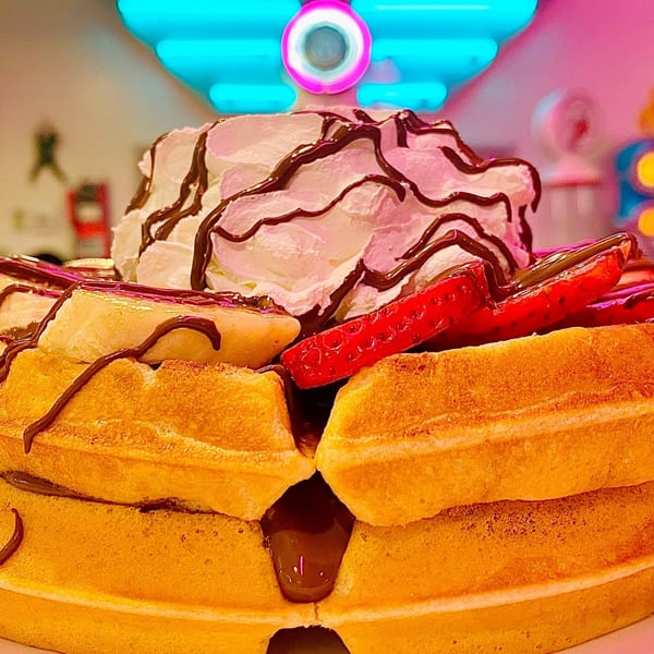Beeps Diner waffles with whipped cream, chocolate drizzle, strawberries, and bananas, under neon lights.